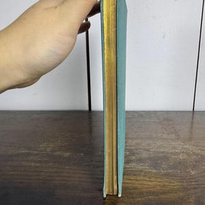 Puede incluir: Un libro de tapa dura de color azul turquesa con bordes dorados. El libro est&aacute; de pie sobre una superficie de madera.