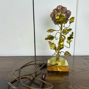 K&ouml;nnte beinhalten: Eine goldfarbene Tischlampe mit einem rosafarbenen Blumendesign. Die Lampe zeigt eine kleine Figur einer Person, die den Stiel der Blume hinaufklettert. Die Lampe hat ein braunes Kabel und einen Schalter.