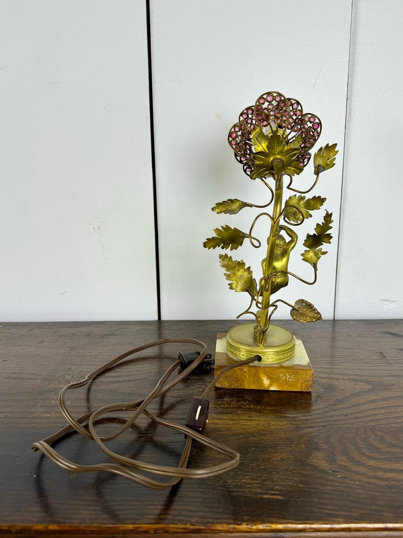 K&ouml;nnte beinhalten: Eine goldfarbene Tischlampe mit einem rosafarbenen Blumendesign. Die Lampe zeigt eine kleine Figur einer Person, die den Stiel der Blume hinaufklettert. Die Lampe hat ein braunes Kabel und einen Schalter.