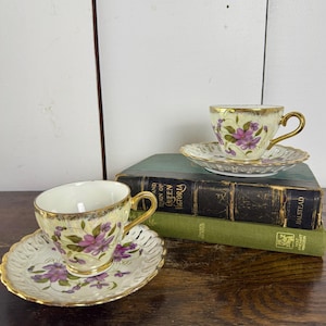 May include: Two antique teacups and saucers with a floral design. The cups and saucers are white with gold trim and feature purple flowers. The saucers have a delicate, openwork design. The teacups are resting on a stack of vintage books.