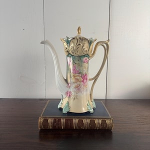May include: A porcelain coffee pot featuring a cream-colored base with a floral pattern of pink and yellow roses. The vessel includes a gold-trimmed handle, a white spout, and a decorative lid with gold accents and light green leaf-like details.
