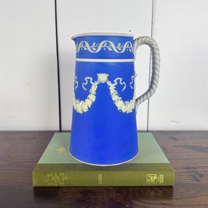 May include: A cobalt blue pitcher with white floral garland and trim. The handle is a twisted rope design. The pitcher is sitting on a green book.