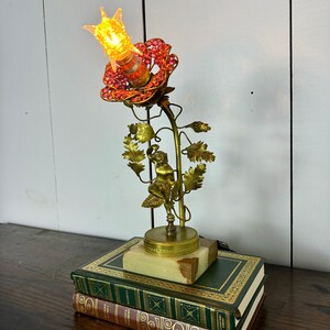 K&ouml;nnte beinhalten: Eine goldene Metalllampe in Form einer Rose mit einer roten, mit Juwelen besetzten Bl&uuml;te und einer klaren Gl&uuml;hbirne. Die Lampe steht auf einem Marmorsockel und ruht auf zwei B&uuml;chern.