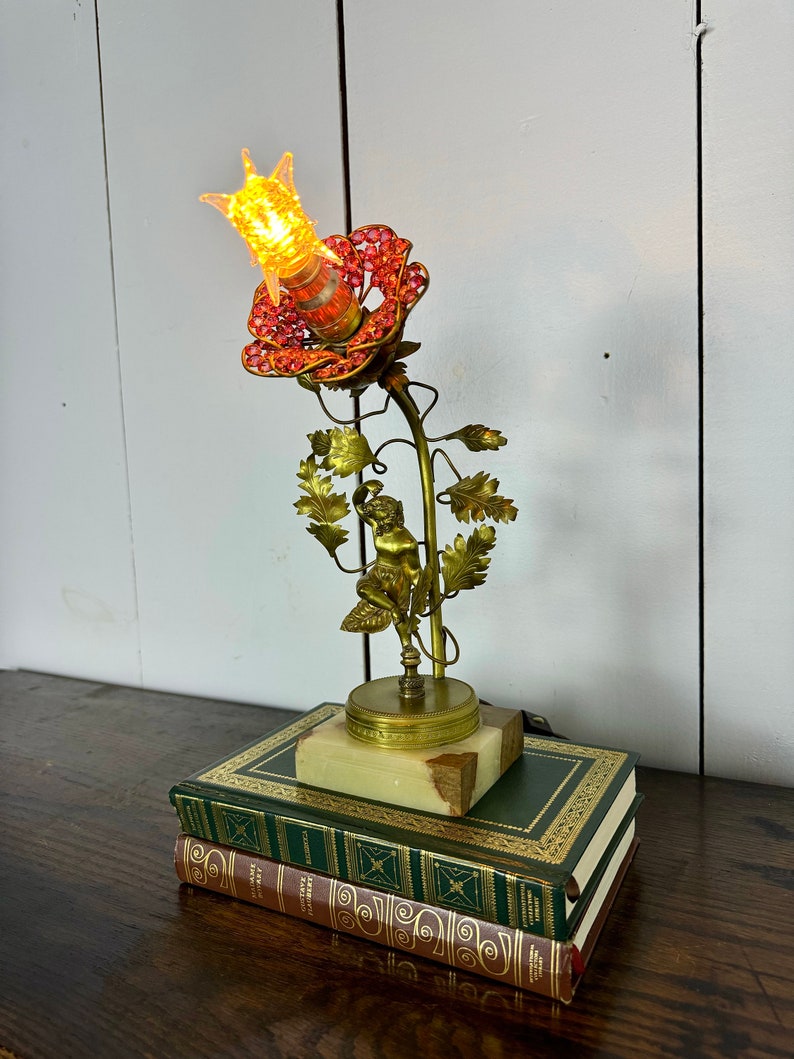 K&ouml;nnte beinhalten: Eine goldene Metalllampe in Form einer Rose mit einer roten, mit Juwelen besetzten Bl&uuml;te und einer klaren Gl&uuml;hbirne. Die Lampe steht auf einem Marmorsockel und ruht auf zwei B&uuml;chern.