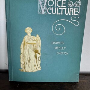 Puede incluir: Un libro azul verdoso con letras doradas que dice "Voice Culture" de Charles Wesley Emerson. La portada presenta una ilustraci&oacute;n dorada de una mujer sosteniendo una lira.