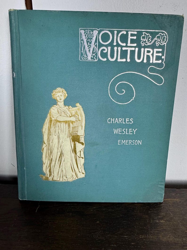 Puede incluir: Un libro azul verdoso con letras doradas que dice "Voice Culture" de Charles Wesley Emerson. La portada presenta una ilustraci&oacute;n dorada de una mujer sosteniendo una lira.