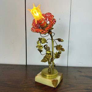 Peut inclure: Une lampe de table en métal doré en forme de fleur avec une figure de chérubin grimpant sur la tige. La fleur est ornée de perles de verre rouges et transparentes et d'une ampoule en verre transparent. La lampe repose sur une base en marbre blanc.