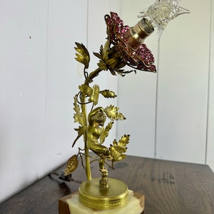K&ouml;nnte beinhalten: Eine goldfarbene Tischlampe mit einem rosafarbenen Schirm, der mit rosa Kristallen verziert ist. Die Lampe zeigt eine Cherubfigur, die an einer Rebe mit Bl&auml;ttern hochklettert. Der Sockel ist aus Marmor.