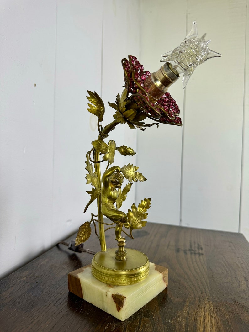 K&ouml;nnte beinhalten: Eine goldfarbene Tischlampe mit einem rosafarbenen Schirm, der mit rosa Kristallen verziert ist. Die Lampe zeigt eine Cherubfigur, die an einer Rebe mit Bl&auml;ttern hochklettert. Der Sockel ist aus Marmor.