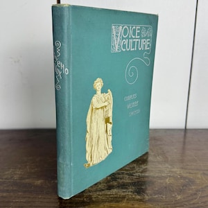 Puede incluir: Un libro de tapa dura azul verdoso titulado "Voice Culture" de Charles Wesley Emerson. La portada presenta una ilustraci&oacute;n dorada de una mujer sosteniendo una lira.