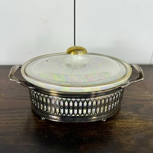 May include: A white porcelain casserole dish with a gold knob on the lid, sitting on a silver metal stand with a decorative design. The dish has a iridescent finish.