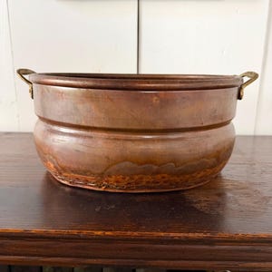 May include: A round copper basin featuring a smooth, polished upper section and a textured, hammered lower base. The vessel includes two small, curved brass handles attached to opposite sides of the rim, showcasing a warm, metallic finish.