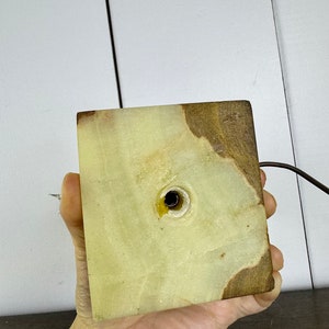 K&ouml;nnte beinhalten: Die Unterseite eines gr&uuml;nen und braunen Alabaster-Lampenfu&szlig;es mit einem Loch f&uuml;r das Kabel. Der Fu&szlig; ist quadratisch und hat eine raue Textur.
