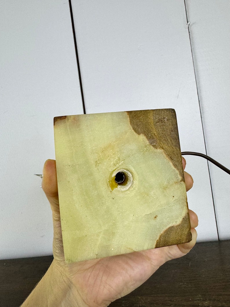 K&ouml;nnte beinhalten: Die Unterseite eines gr&uuml;nen und braunen Alabaster-Lampenfu&szlig;es mit einem Loch f&uuml;r das Kabel. Der Fu&szlig; ist quadratisch und hat eine raue Textur.
