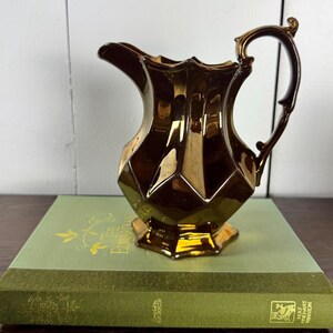 May include: A gold-colored ceramic pitcher with a faceted body and a decorative handle. The pitcher is sitting on a green book with gold lettering.