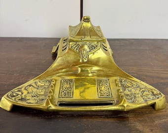 Peut inclure: Un encrier en laiton vintage avec un design Art nouveau. L'encrier présente des motifs floraux et géométriques, avec un compartiment central et un couvercle décoratif. Le laiton a une patine chaude et vieillie, et la pièce est posée sur une surface en bois sombre.