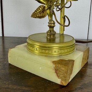 K&ouml;nnte beinhalten: Eine goldene Cherubstatue mit einem Blattdesign, die auf einem runden Sockel mit einem dekorativen Rand steht. Der Sockel ist aus gr&uuml;nem Marmor und steht auf einer Holzoberfl&auml;che.