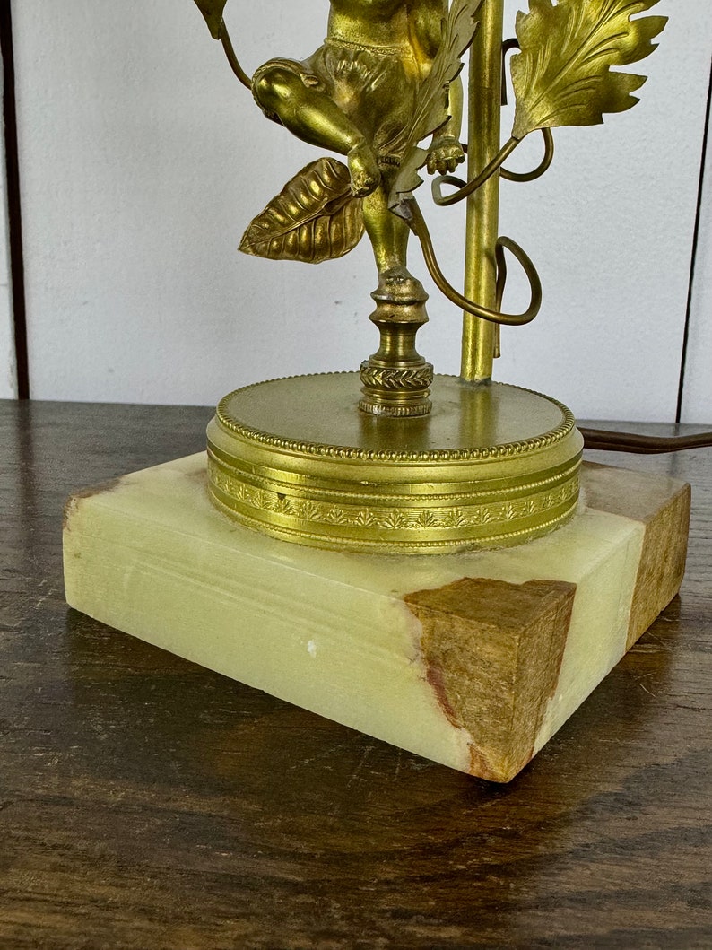 K&ouml;nnte beinhalten: Eine goldene Cherubstatue mit einem Blattdesign, die auf einem runden Sockel mit einem dekorativen Rand steht. Der Sockel ist aus gr&uuml;nem Marmor und steht auf einer Holzoberfl&auml;che.