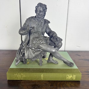 May include: A detailed pewter sculpture of a seated man in period clothing, resting on a green book. The figure is holding a book and is seated on a small chair. The book is approximately 8 inches long.