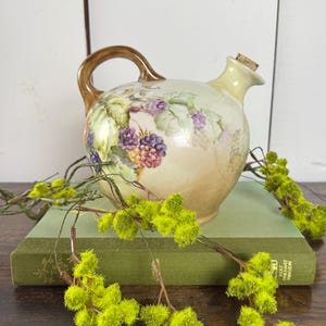 May include: A vintage ceramic pitcher with a brown handle and spout, featuring a hand-painted design of grapes and leaves in shades of purple, green, and cream. The pitcher has a cork stopper and is resting on a green book, with yellow floral accents.