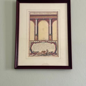 May include: Framed vintage architectural print with a burgundy wooden frame. The print features a detailed illustration of a classical building facade with arches, columns, and decorative elements. The print has text in French.