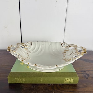 May include: A white porcelain serving dish with a scalloped edge and gold trim. The dish is oval-shaped and sits atop a green book. The dish has ornate detailing and a decorative design.