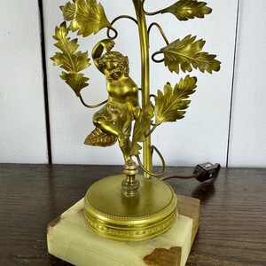 K&ouml;nnte beinhalten: Eine goldfarbene Tischlampe mit einer Skulptur einer Frau, die an einer Weinrebe mit Bl&auml;ttern hochklettert. Die Lampe hat einen gr&uuml;nen Marmorsockel.