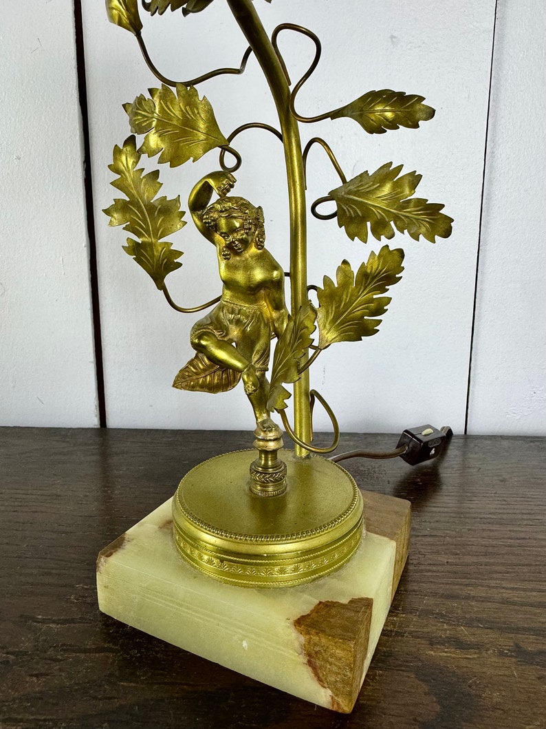 K&ouml;nnte beinhalten: Eine goldfarbene Tischlampe mit einer Skulptur einer Frau, die an einer Weinrebe mit Bl&auml;ttern hochklettert. Die Lampe hat einen gr&uuml;nen Marmorsockel.