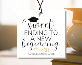 Puede incluir: Una etiqueta cuadrada blanca con texto negro que dice "A sweet ending to a new beginning" con un icono de gorro de graduación sobre el texto. El texto "Congratulations Grad!" está debajo del texto principal. La etiqueta está colgada de una cuerda negra.