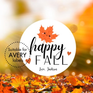 Puede incluir: Una pegatina circular blanca con el texto "happy fall" en fuente cursiva negra. Una hoja de arce naranja de dibujos animados con una cara sonriente está por encima del texto. El texto "Love: Jackson" está debajo del texto "happy fall". La pegatina está sobre un fondo de hojas de otoño borrosas de color naranja y amarillo.