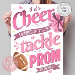 Puede incluir: Un póster rosa y blanco con el texto "I'd Cheer So Loud If You'd Tackle Prom With Me!". También se incluye un balón de fútbol americano y un pompón. El póster tiene un estilo de acuarela y lo sostiene una persona.