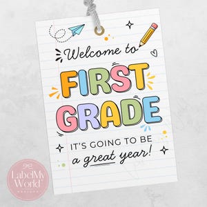 May include: A rectangular paper tag with a grey string featuring a lined notebook paper background. The text reads Welcome to FIRST GRADE IT'S GOING TO BE a great year! with colourful bubble letters, a yellow pencil, and a blue paper aeroplane.