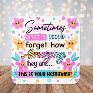 May include: A white square sign with a pink, yellow, and blue floral design. The sign has the text "Sometimes amazing people forget how Amazing they are... This is your Reminder!"