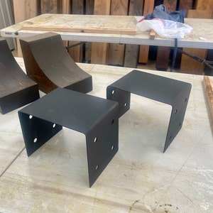 Może przedstawiać: Dwa czarne metalowe wsporniki i dwa drewniane wsporniki. Metalowe wsporniki są prostokątne ze zaokrąglonymi narożnikami i mają po cztery otwory z każdej strony. Drewniane wsporniki są zakrzywione i mają ciemnobrązowe wykończenie.