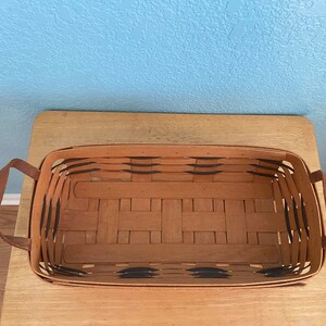 May include: A rectangular woven basket with black accents and two brown leather handles. The basket is empty and is sitting on a wooden surface.