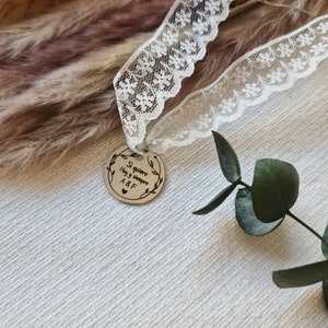 Puede incluir: Un charm redondo plateado con el texto "Si quiero hoy y siempre A&F" grabado en él. El charm está unido a una cinta de encaje blanco.
