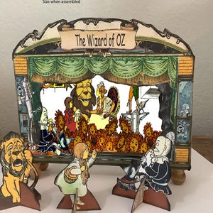 May include: A miniature Wizard of Oz theater set with a stage, characters, and the text "The Wizard of Oz." The stage has a green curtain, and the characters include Dorothy, the Lion, and the Tin Man. The set is made of cardboard.