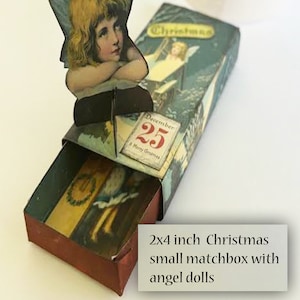 May include: A 2x4 inch Christmas matchbox with a pop-up angel doll. The matchbox is decorated with a blue and white winter scene and the text "Christmas". The date "December 25" is visible on the front of the matchbox.