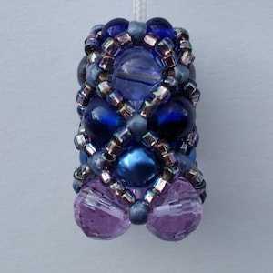 Może przedstawiać: Ciągnąca lampę z koralikami w kolorze niebieskim i fioletowym. Ciągnąca jest wykonana z małych szklanych koralików i ma geometryczny wzór.