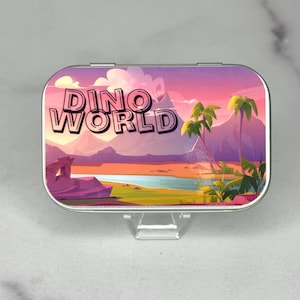 Puede incluir: Una lata metálica rectangular con una colorida ilustración de un paisaje y el texto "DINO WORLD". La obra de arte presenta un cielo al atardecer, montañas, palmeras y un cuerpo de agua.