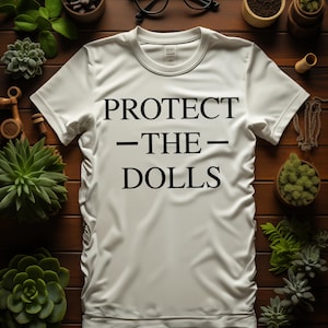 May include: White t-shirt with the text "PROTECT - THE - DOLLS" in black. The shirt is on a dark wood surface, surrounded by small potted succulents and a pair of black eyeglasses.