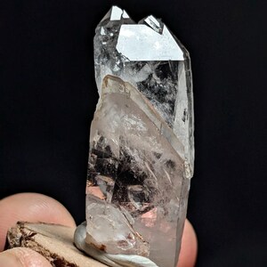 May include: A clear, prismatic quartz crystal specimen featuring a sharp, pointed termination and internal fractures. The crystal is mounted upright on a small, circular, textured wooden base, showcasing natural transparency and geometric facets.