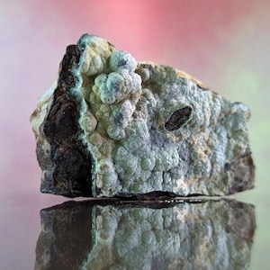 May include: A close-up of a mineral specimen with a textured, bubbly surface. The rock has a mix of colors, including light blue, gray, and dark brown. The mineral is reflected in the surface below, with a soft pink and green background.