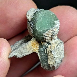 May include: A green crystal cluster with a rough, gray exterior. The crystal is partially embedded in a gray rock matrix.