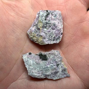 May include: Two pieces of raw, natural mineral specimens. The stones have a mix of colors, including lavender, gray, and black. The rough texture and irregular shapes suggest they are unpolished and in their natural state.