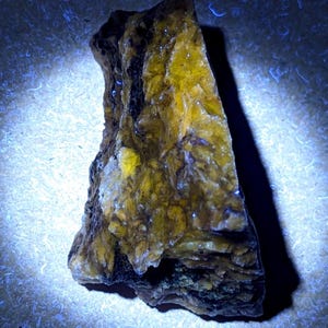 May include: A rough-hewn, irregularly shaped mineral specimen. The rock has a layered appearance with a mix of yellow, brown, and black hues. The surface has a crystalline texture, and the lighting creates highlights and shadows.