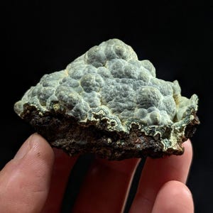 May include: A close-up of a mineral specimen held in a hand. The mineral has a bumpy, light blue-gray surface with a dark brown base. The edges of the mineral have a white and green coloration.
