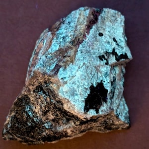 May include: A rough-hewn rock with a mix of colors, including black, gray, and patches of light blue. The rock has a textured surface and irregular shape, with a dark, almost black, area on one side. The overall appearance suggests a natural mineral specimen.
