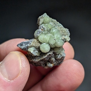 May include: A small rock with green, ball-shaped crystals. The crystals are clustered together on the rock, which is gray and brown.