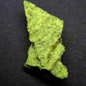 May include: A vibrant, irregularly shaped mineral specimen with a striking, bright yellow-green color. The surface has a textured, crystalline appearance. The background is a dark, textured surface, providing contrast to the glowing mineral.
